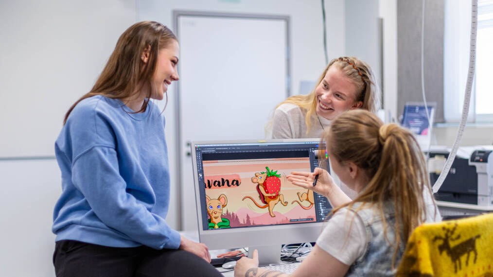 Kolme nuorta henkilöä työskentelee yhdessä graafisen suunnittelun projektin parissa tietokoneella. Näytöllä näkyy piirros hiirestä ja mansikasta.