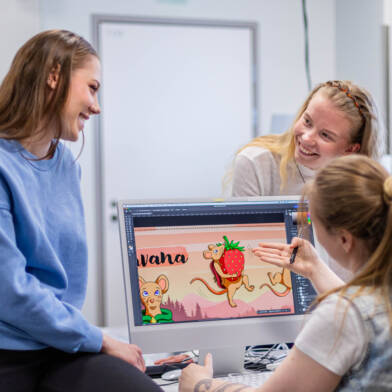 Kolme nuorta henkilöä työskentelee yhdessä graafisen suunnittelun projektin parissa tietokoneella. Näytöllä näkyy piirros hiirestä ja mansikasta.