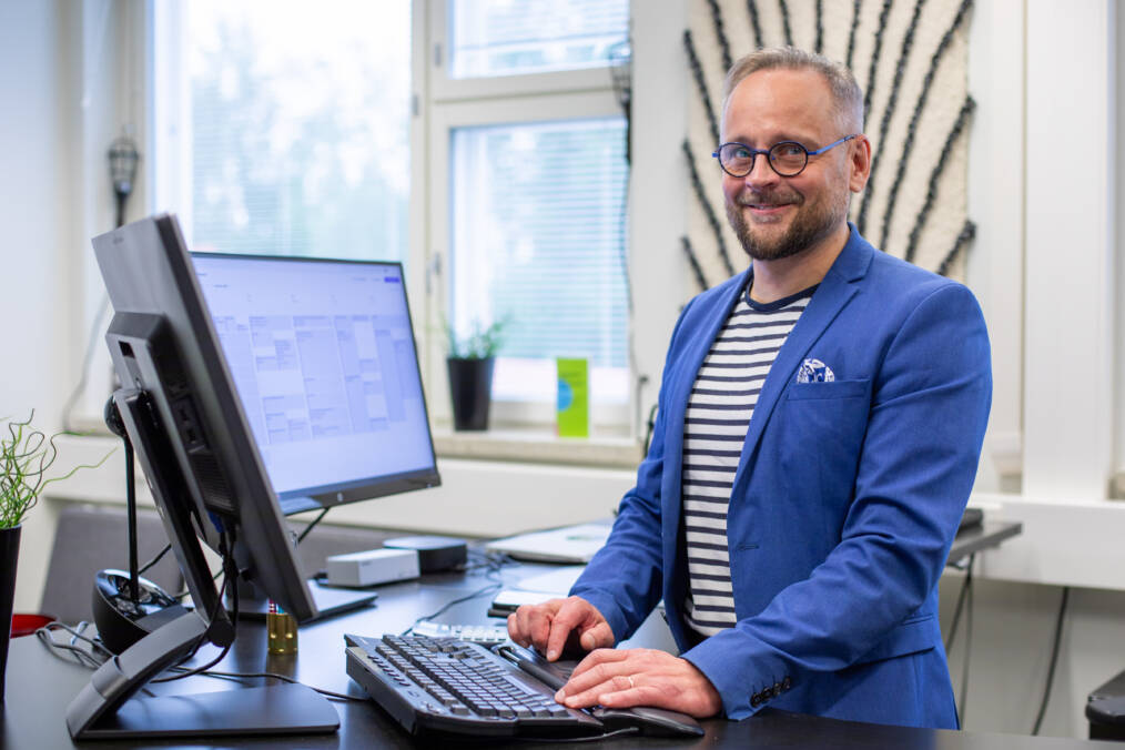 Henkilö työskentelemässä tietokoneella ikkunan vieressä sijaitsevalla työpisteellä, henkilö katsoo kameraan ja hymyilee.