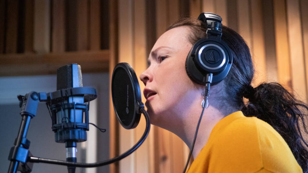 Henkilö laulaa mikrofoniin äänitysstudiossa kuulokkeet päässään.