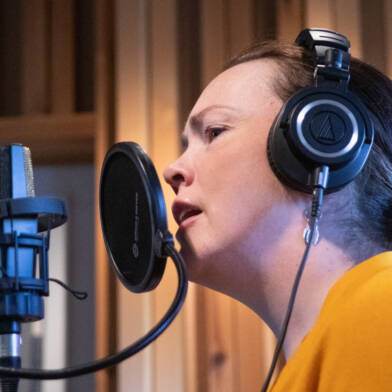Henkilö laulaa mikrofoniin äänitysstudiossa kuulokkeet päässään.