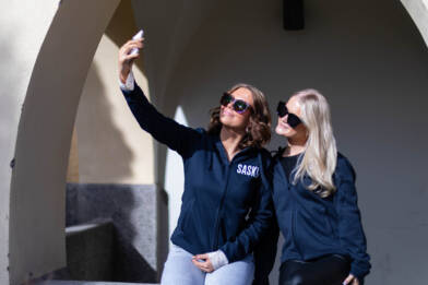 Kaksi nuorta henkilöä ottamassa selfietä auringonpaisteessa, päällään SASKY-hupparit.