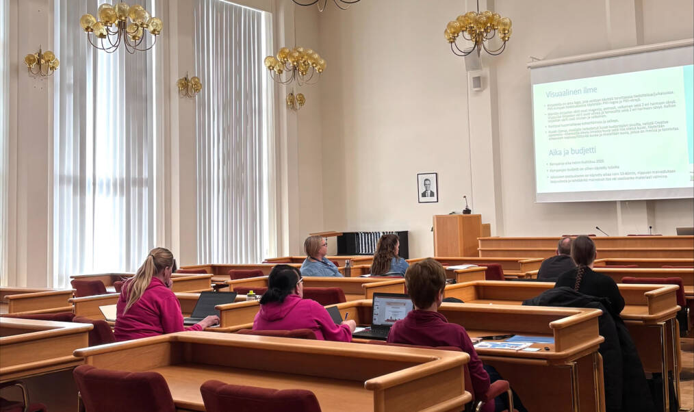 Ryhmä henkilöitä koulutustilanteessa isossa kokoussalissa tai vastaavassa tilassa, tilan katosta roikkuu näyttäviä valaisimia.