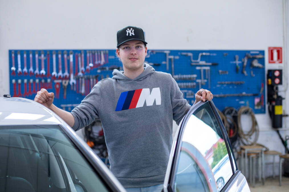 Harmaaseen huppariin ja mustaan lippikseen pukeutunut autoalan opiskelija auton vieressä hallissa, taustalla näkyy sinisellä pohjalla oleva työkaluseinä.