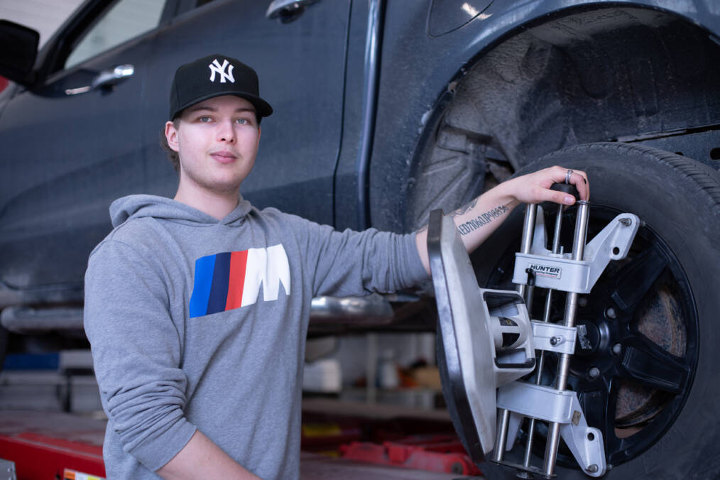 Harmaaseen huppariin ja mustaan lippikseen pukeutunut autoalan opiskelija nosturilla olevan auton takarenkaan vieressä, auton renkaassa kiinni mittauslaite.