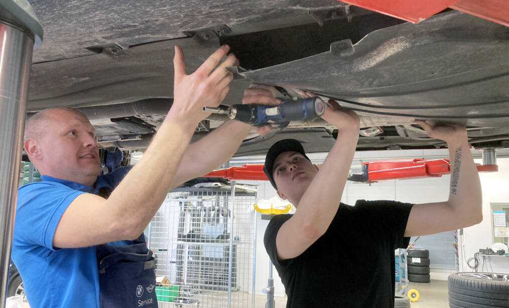 Autoalan opiskelija työskentelee auton pohjan parissa nosturilla, mukana myös tilannetta ohjaava henkilö.