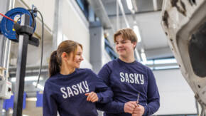 Kaksi henkilöä lyö leikkisästi kyynärpäitään yhteen. Molemmilla sininen pusero, jossa tekstilogo SASKY