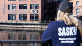 Kuvassa vettä ja taustalla punatiilinen rakennus, jossa Saskyn logo. Kuvan etualalla sumeana henkilö takaapäin nojaamassa kaiteeseen.
