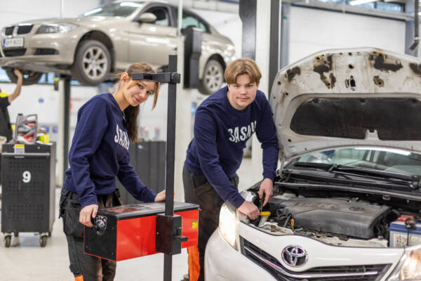 Kaksi autoalan opiskelijaa työskentelee autokorjaamossa auton konepellin äärellä, taustalla auto nosturilla.