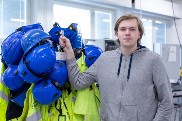 Harmaaseen huppariin pukeutunut henkilö seisoo sisätilassa ja pitää kädellään telineestä, jossa roikkuu sinisiä kypäriä ja neonkeltaisia huomioliivejä.