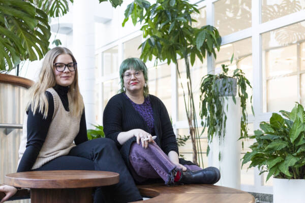 Kaksi henkilöä istuu sisätilassa viherkasvien ympäröimänä ja katsoo kameraan; taustalla on valoisa aula tai käytävä.