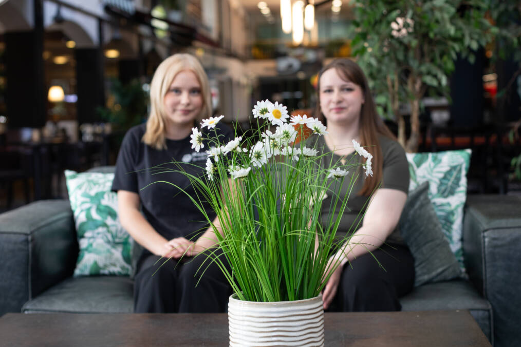 Kaksi henkilöä istuu vierekkäin sohvalla, taustalla näkyy kylpylä-hotellin aulatilaa, tarkennus on kohdistettu kuvan etualalla pöydällä olevaan kukka-asetelmaan.