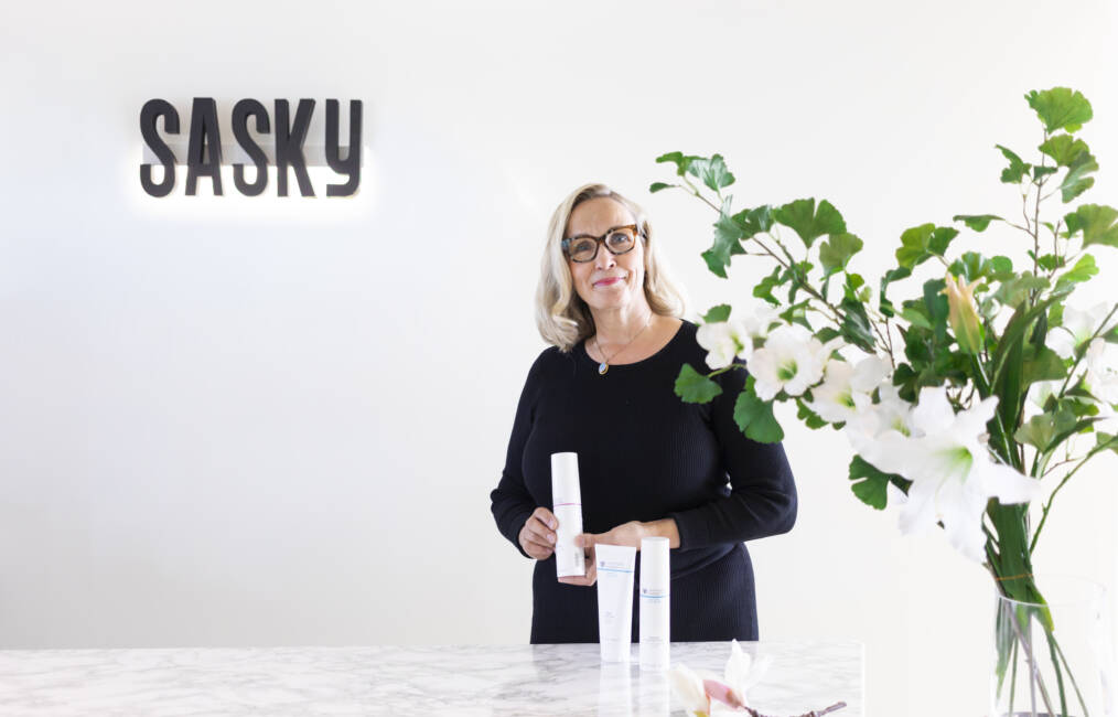 Vaaleahiuksinen silmälasipäinen henkilö seisoo valoisassa tilassa SASKY-logon edessä ja esittelee ihonhoitotuotteita. Etualalla näkyy kukkia maljakossa.