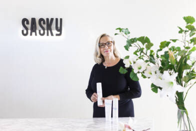 Vaaleahiuksinen silmälasipäinen henkilö seisoo valoisassa tilassa SASKY-logon edessä ja esittelee ihonhoitotuotteita. Etualalla näkyy kukkia maljakossa.