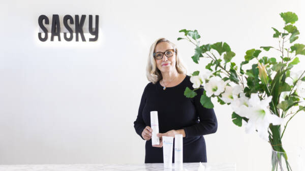 Vaaleahiuksinen silmälasipäinen henkilö seisoo valoisassa tilassa SASKY-logon edessä ja esittelee ihonhoitotuotteita. Etualalla näkyy kukkia maljakossa.