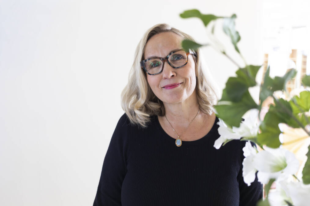 Lähikuva hymyilevästä vaaleahiuksisesta silmälasipäisestä henkilöstä valoisassa hoitolaympäristössä, valkoisia kukkia etualalla.