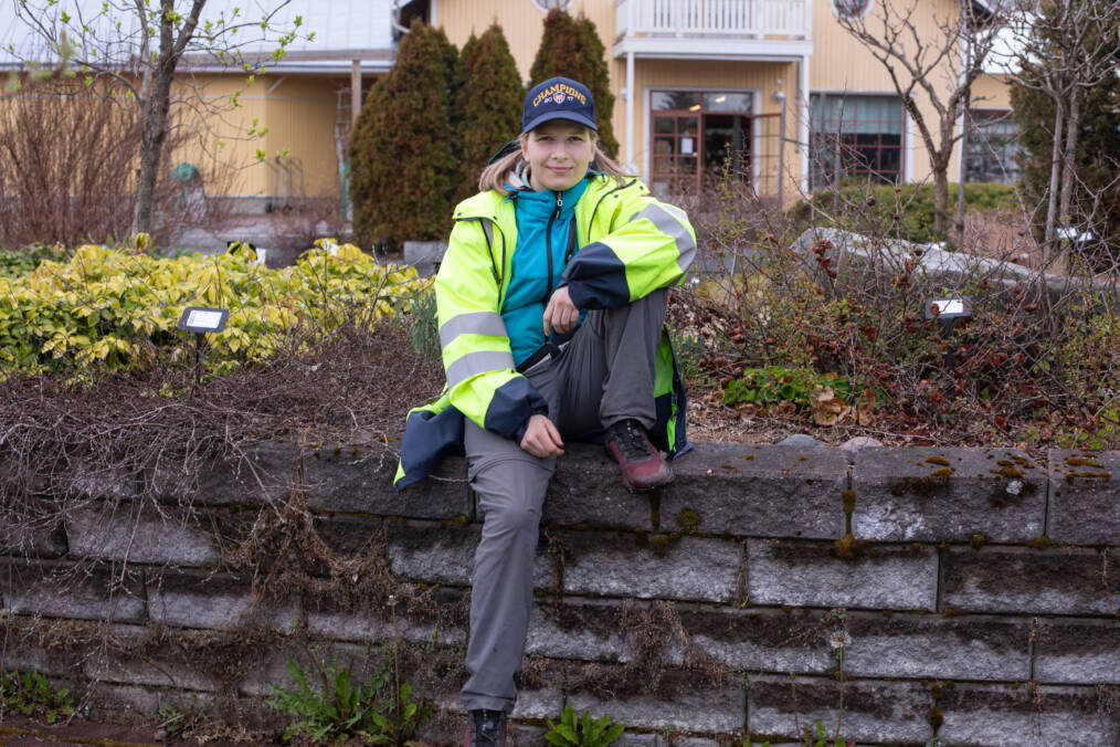Henkilö istuu kivimuurin päällä puutarha-alueella, taustalla kasveja ja rakennus.