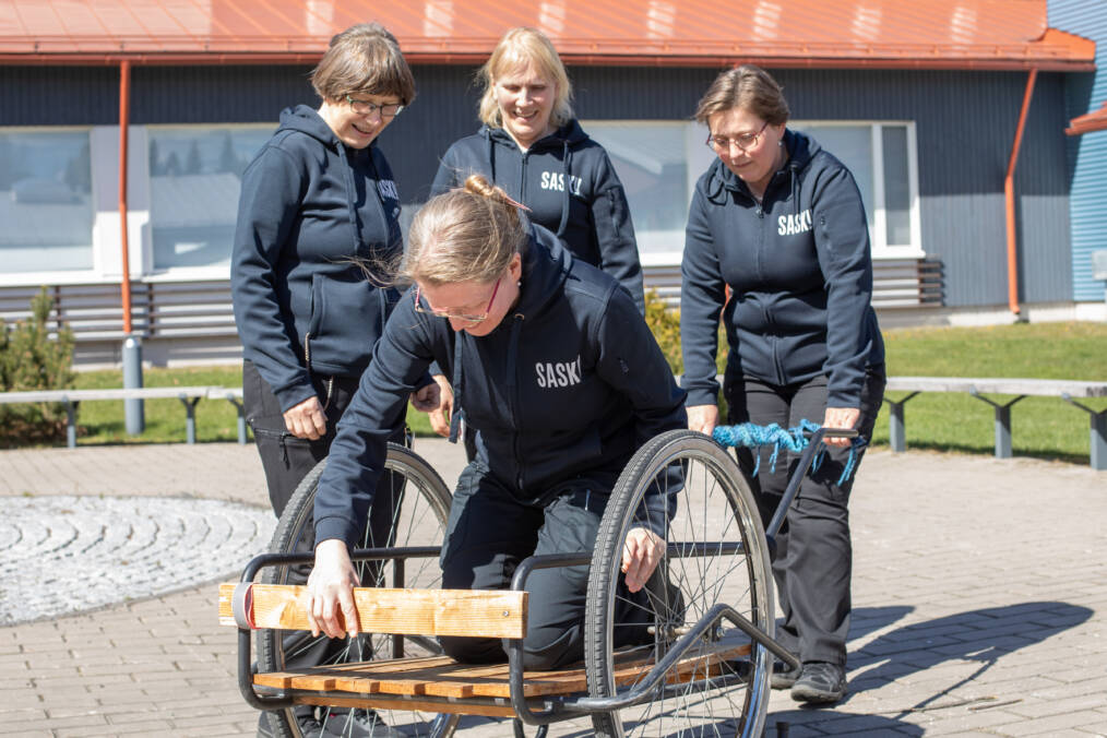 Yksi henkilö on menossa istumaan maitokärryhin ja toinen henkilö pitää kiinni kärryn aisasta, kaksi henkilöä seisoo heidän vieressään pihalla, henkilöillä on päällään tummansiniset hupparit, joissa valkoisella teksti Sasky.