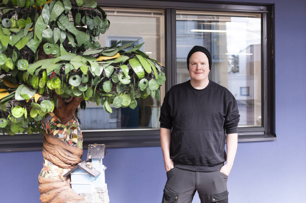Musta pipo ja collegepaita päällä oleva henkilö seisoo ulkona ikkunan edessä suuren koristeellisen puuveistoksen vieressä.
