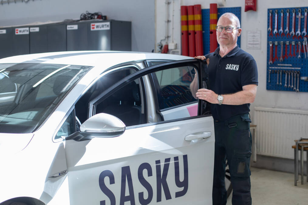 Opettaja seisoo auton vieressä autokorjaamossa, hänellä on päällään tummansininen paita, jossa valkoisella Sasky-teksti, valkoisen auton kyljessä tummansinisellä Sasky-teksti.