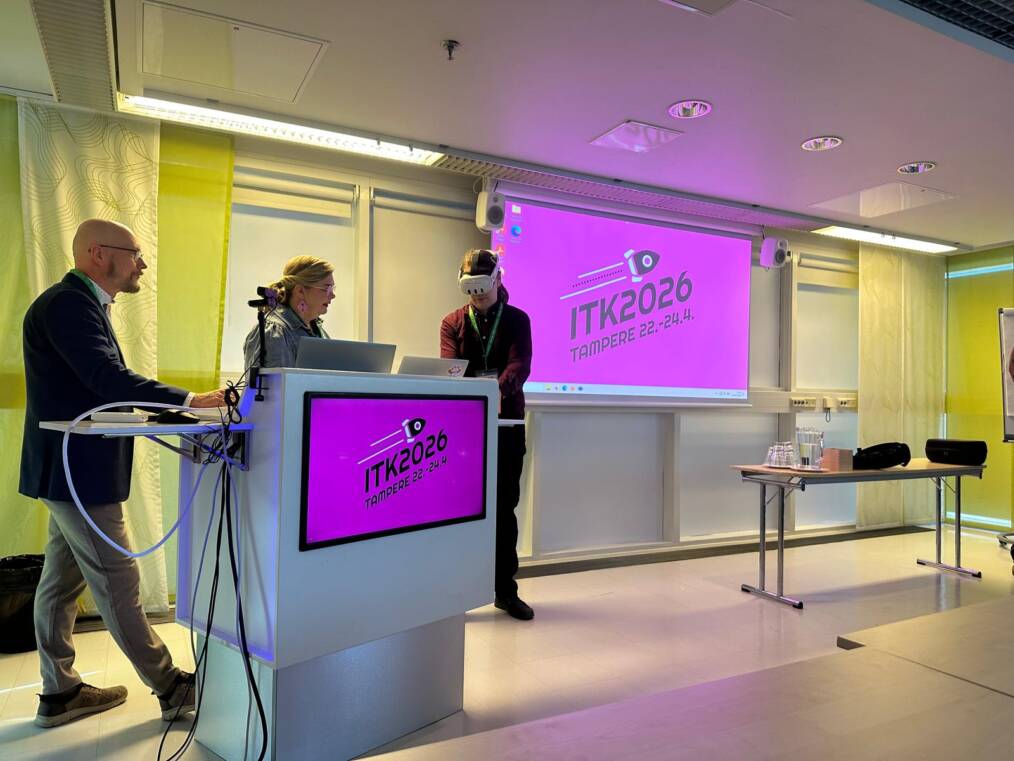 Koulutustilanteessa kaksi henkilöä esittelee VR-teknologiaa yleisölle ITK2026-tapahtumassa. Yksi osallistuja käyttää VR-laseja puhujakorokkeen vieressä, samalla kun taustalla näkyy tapahtuman pinkki ITK2026-esitysnäkymä.