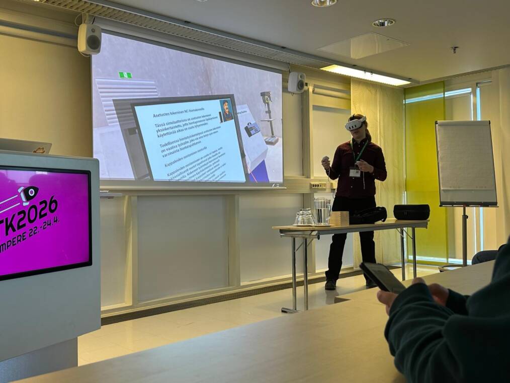 Henkilö esittelee VR-simulaatiota koulutustilassa käyttäen VR-laseja. Taustalla valkokankaalla näkyy virtuaalinen oppimisympäristö ja ITK2026-tapahtuman visuaalinen ilme.