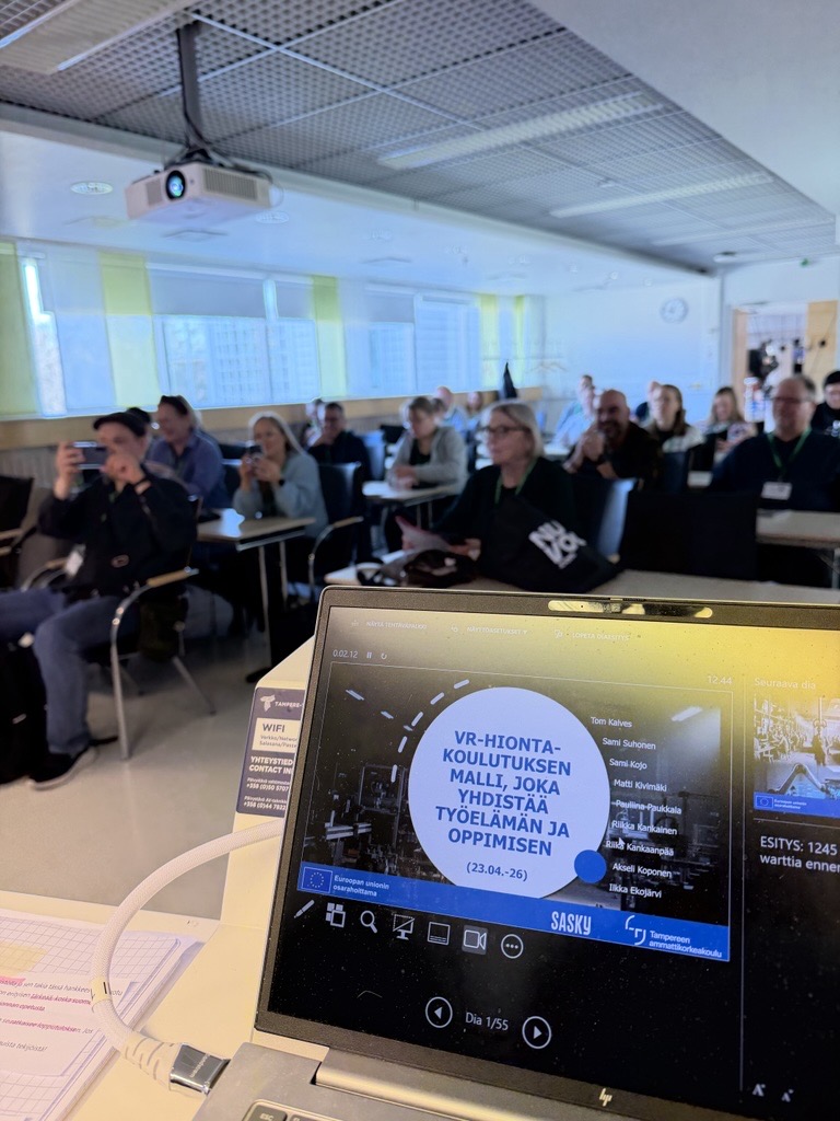 Yleisö seuraa VR-hiontakoulutuksen esitystä luokkatilassa. Etualalla näkyy kannettava tietokone, jonka näytöllä on Saskyn ja Tampereen ammattikorkeakoulun yhteinen esitysdia aiheesta ”VR-hiontakoulutuksen malli, joka yhdistää työelämän ja oppimisen”.