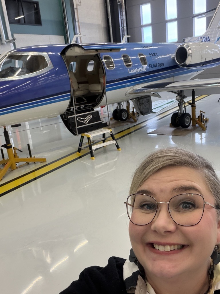 Hymyilevä henkilö ottaa selfien lentokonehallissa sinivalkoisen Learjet 24D -suihkukoneen edessä. Koneen ovi on auki ja portaat ovat paikallaan huoltotilan lattialla.