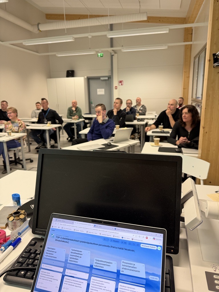 Koulutustilanne luokkahuoneessa, jossa osallistujat istuvat pöytien ääressä ja seuraavat esitystä. Etualalla näkyy tietokoneen näyttö sekä kannettava, jonka ruudulla on työelämäpalautteen kehittämiskohteita käsittelevä näkymä.