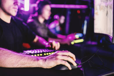 Esports-pelaajia tietokoneilla violetiksi valaistussa pelitilassa. Etualalla pelaajan käsi tietokonehiirellä.