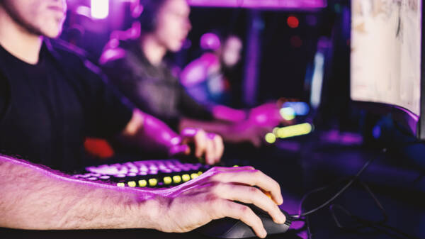 Esports-pelaajia tietokoneilla violetiksi valaistussa pelitilassa. Etualalla pelaajan käsi tietokonehiirellä.