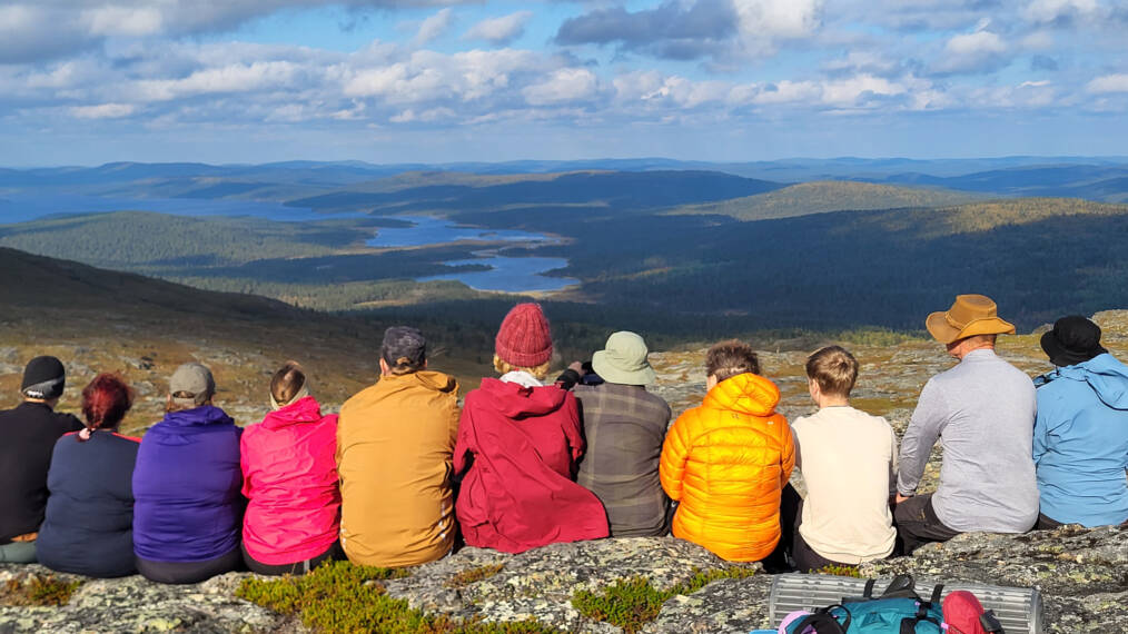 Ryhmä henkilöitä istuu rivissä tunturin laella ja on selin kameraan, taustalla näkyy tunturimaisemaa järvineen.