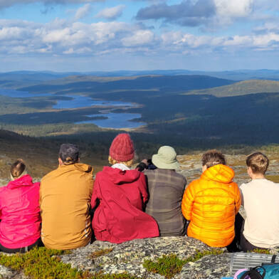 Ryhmä henkilöitä istuu rivissä tunturin laella ja on selin kameraan, taustalla näkyy tunturimaisemaa järvineen.