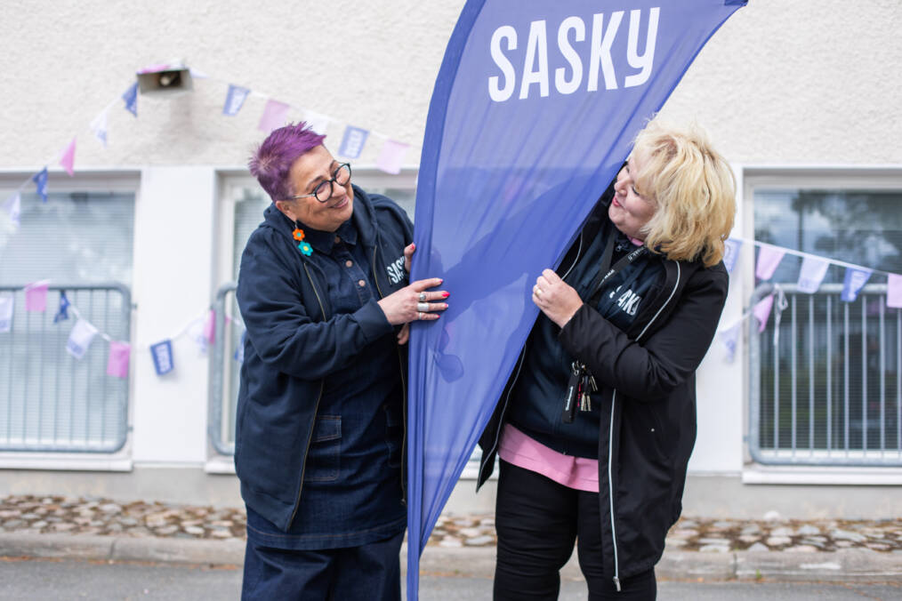 Kaksi henkilöä seisoo Saskyn lipun vieressä ulkoalueella. Taustalla näkyy rakennus ja viirejä.