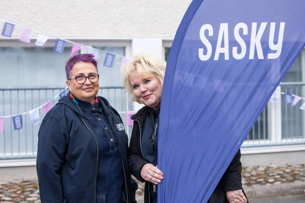 Kaksi henkilöä seisoo Saskyn lipun vieressä ulkoalueella. Taustalla näkyy rakennus ja viirejä.