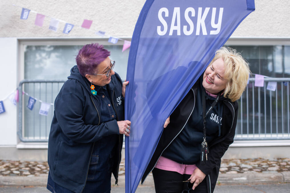 Kaksi henkilöä seisoo Saskyn lipun vieressä ulkoalueella. Taustalla näkyy rakennus ja viirejä.