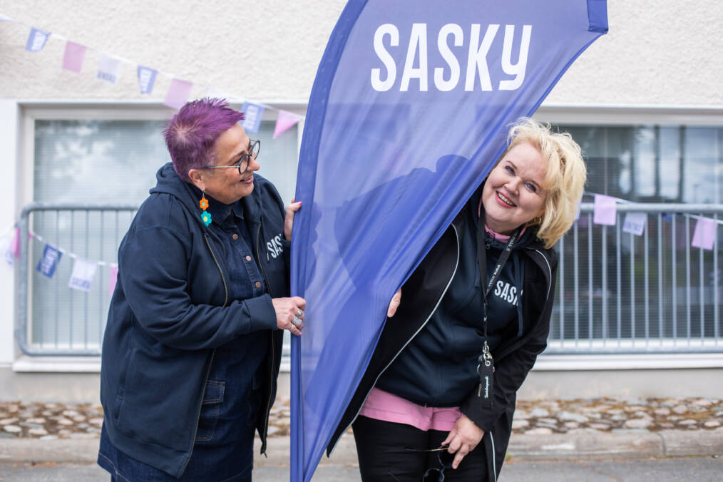Kaksi henkilöä seisoo Saskyn lipun vieressä ulkoalueella. Taustalla näkyy rakennus ja viirejä.