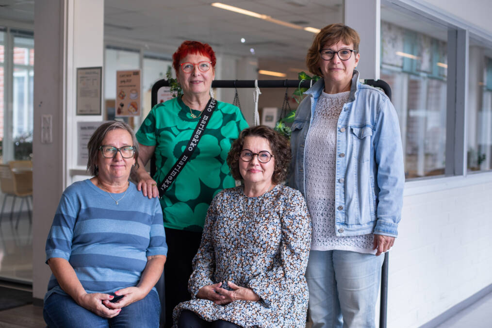 Neljä henkilöä seisoo ja istuu ryhmäkuvassa oppilaitoksen käytävätilassa. Taustalla näkyy sisätiloja ja kasveilla koristeltu teline.