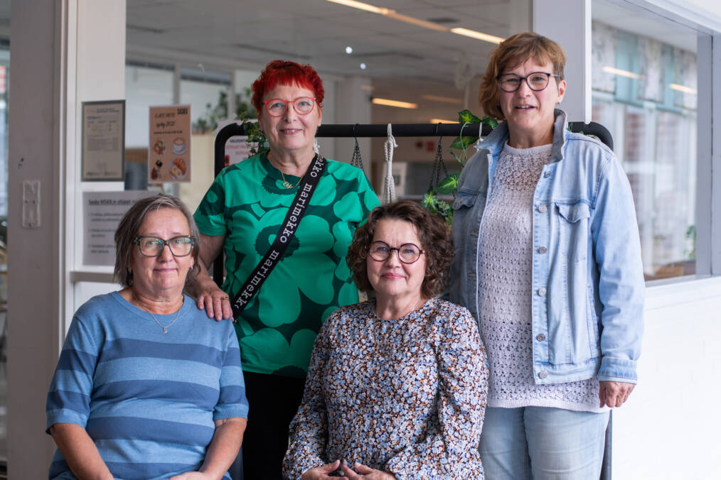 Neljä henkilöä seisoo ja istuu ryhmäkuvassa oppilaitoksen käytävätilassa. Taustalla näkyy sisätiloja ja kasveilla koristeltu teline.