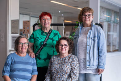 Neljä henkilöä seisoo ja istuu ryhmäkuvassa oppilaitoksen käytävätilassa. Taustalla näkyy sisätiloja ja kasveilla koristeltu teline.