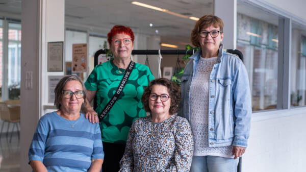 Neljä henkilöä seisoo ja istuu ryhmäkuvassa oppilaitoksen käytävätilassa. Taustalla näkyy sisätiloja ja kasveilla koristeltu teline.