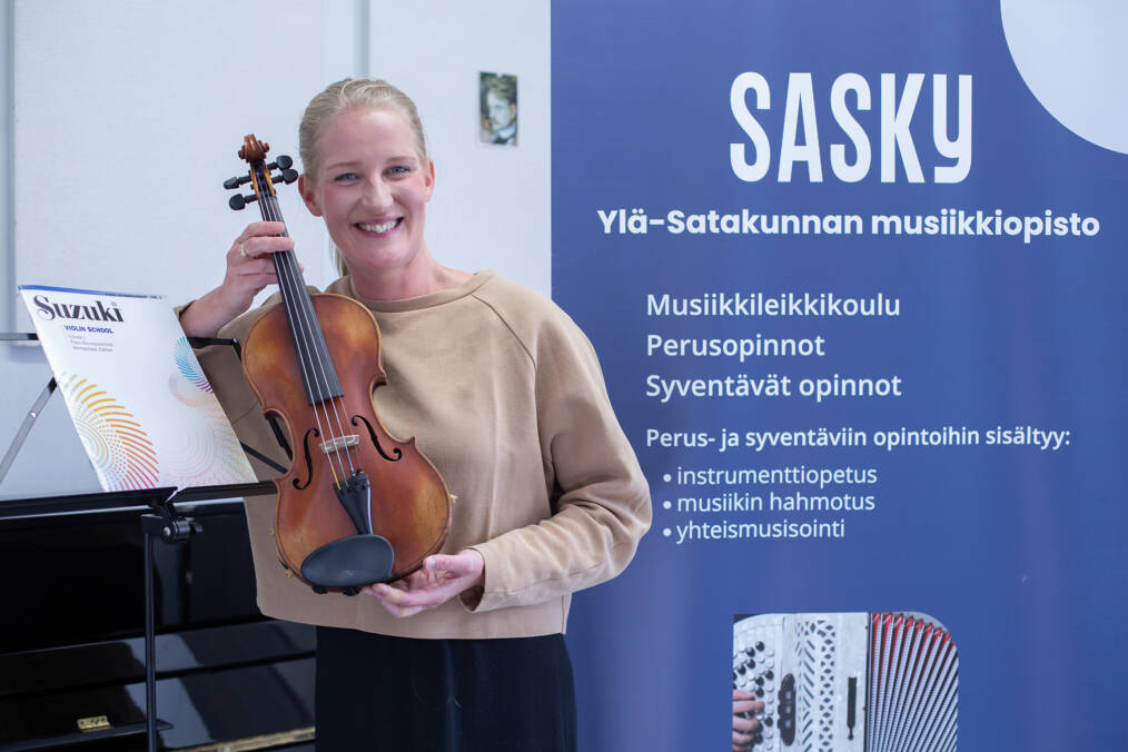 Vaaleanruskeaan paitaan pukeutunut hymyilevä henkilö, jolla on viulu kädessään, vieressä nuottiteline, jossa Suzuki -niminen nuottikirja ja taustalla juliste, jossa on tietoa Sasky Ylä-Satakunnan musiikkiopistosta.