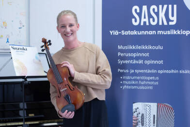Vaaleanruskeaan paitaan pukeutunut hymyilevä henkilö, jolla on viulu kädessään, vieressä nuottiteline, jossa Suzuki -niminen nuottikirja ja taustalla juliste, jossa on tietoa Sasky Ylä-Satakunnan musiikkiopistosta.