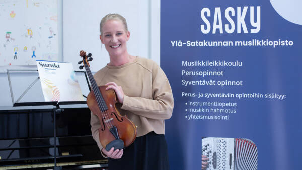 Vaaleanruskeaan paitaan pukeutunut hymyilevä henkilö, jolla on viulu kädessään, vieressä nuottiteline, jossa Suzuki -niminen nuottikirja ja taustalla juliste, jossa on tietoa Sasky Ylä-Satakunnan musiikkiopistosta.