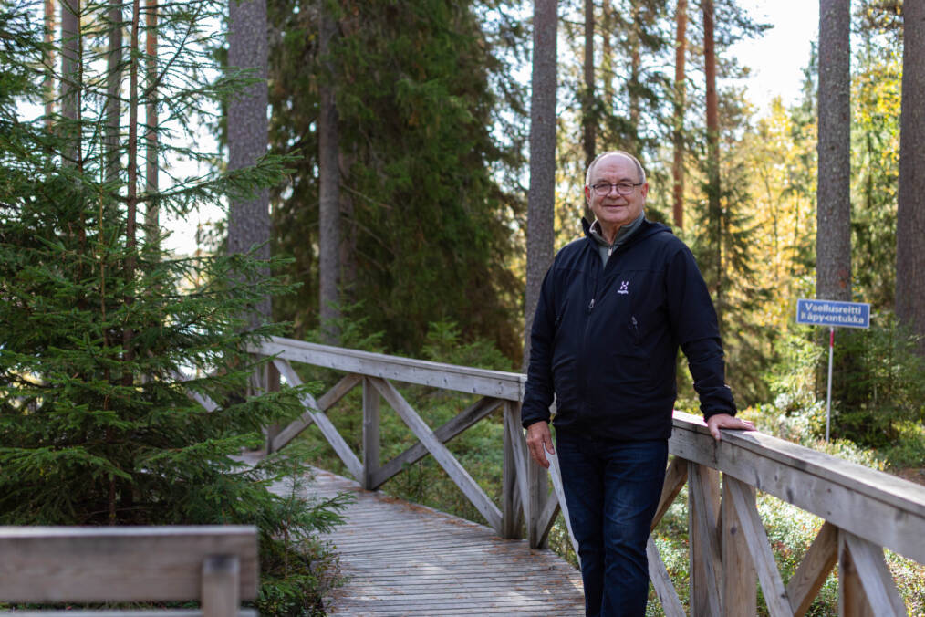 Hannu Raitio seisoo metsäisessä ympäristössä pitkospuureitin kaiteen vieressä.