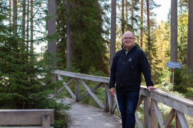 Hannu Raitio seisoo metsäisessä ympäristössä pitkospuureitin kaiteen vieressä.