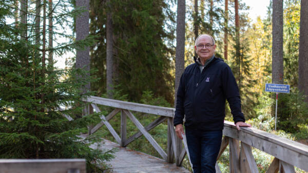 Hannu Raitio seisoo metsäisessä ympäristössä pitkospuureitin kaiteen vieressä.