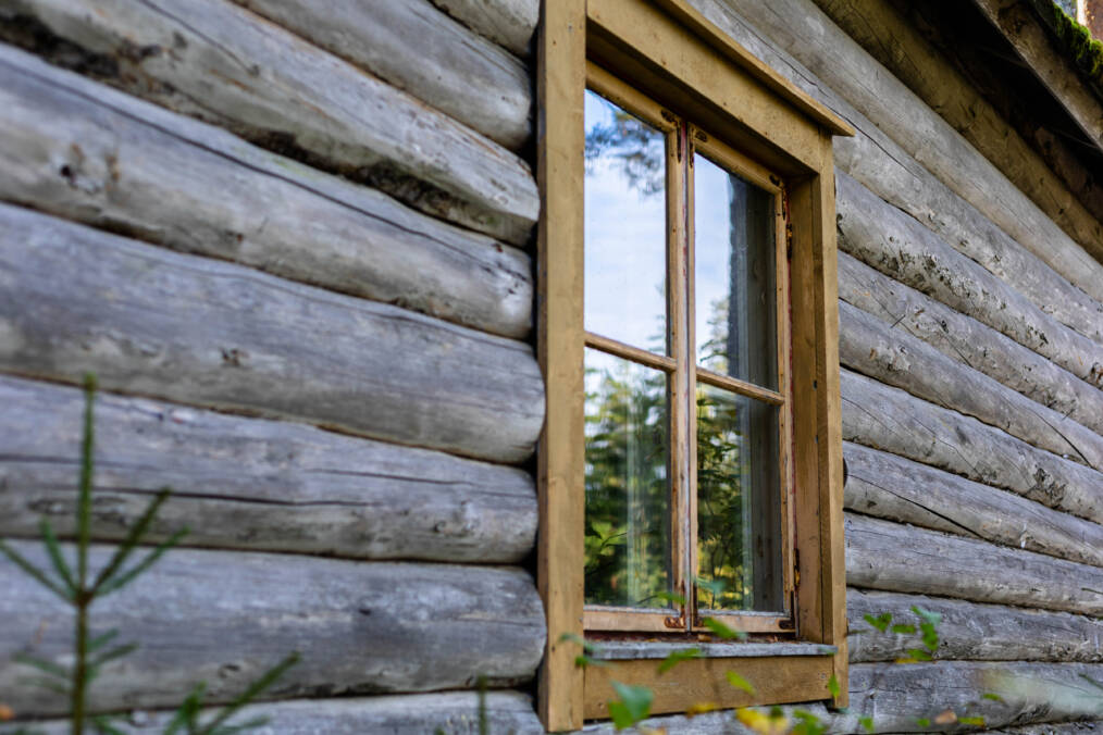 Vanhan hirsirakennuksen ikkuna ja harmaantunutta hirsiseinää metsäisessä ympäristössä.